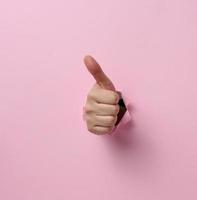 la mano de una mujer sobresale del agujero rasgado y muestra un gesto bien, pulgar arriba. fondo rosa foto