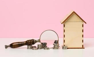 stack of metallic money and magnifier and wooden house on a pink background. Real estate rental photo