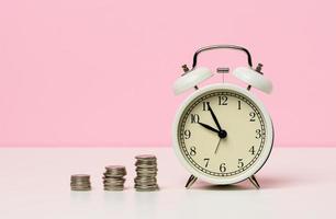 stack of coins round white alarm clock on the table. The concept of time is money, a period of discounts. photo