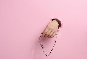 female hand holding glasses, part of body sticking out of torn hole in pink paper background photo