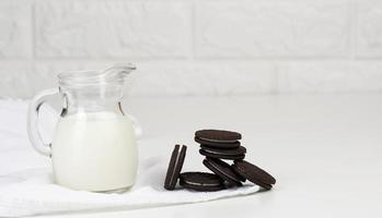 full glass carafe of milk and a stack of chocolate chip cookies on a white table photo