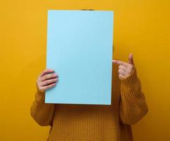 woman in an orange sweater holds a blank sheet of paper on a yellow background. Place for an inscription, advertisement, information photo