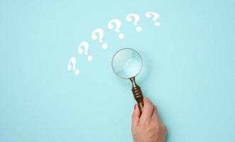 female hand holds a plastic magnifying glass and question marks on a blue background. The concept of finding an answer to questions, truth and uncertainty. photo