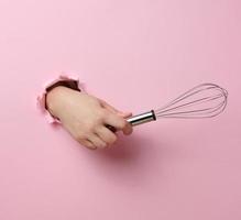 female hand holds a metal whisk for beating products on a pink background photo