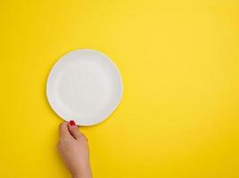 female hand holding a white empty round plate on a yellow background, top view photo