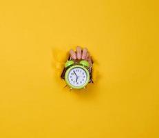 a woman's hand holds a round green alarm clock, the time is five minutes to seven in the morning. A part of the body is sticking out of a torn hole in a yellow paper background. photo