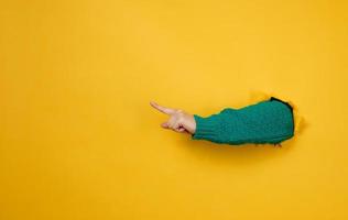 la mano femenina sobresale de un agujero rasgado en un fondo de papel amarillo, parte del cuerpo apunta con el dedo índice hacia un lado. copie el espacio foto