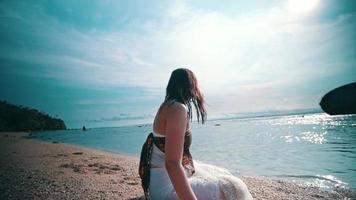 ein asiatisch Frau mit nass blond Haar ist Sitzung auf das Strand während genießen das Aussicht von das Blau Meer video