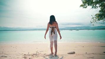 an Asian woman with blonde hair standing on the beach while enjoying the view of the blue sea water and white sand video