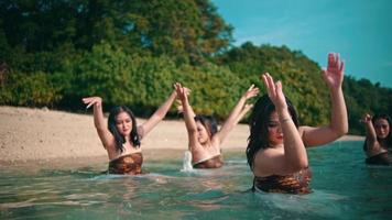 a group of asian women in brown clothes dancing together in the blue sea water on vacation on an island full of green trees during video