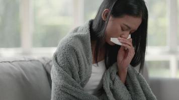 Footage of unhappy sad Asian woman in cloak suffering from fever and flu on sofa blow your nose with a napkin in the living room. Health care and medical concepts. video