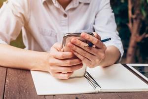 asian woman hand holding phone and pencil for writing notebook in coffee shop with vintage toned photo