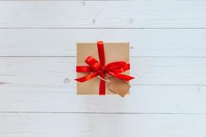 Above brown gift box with tag on wooden board background. Gift box with red ribbon on wooden white background with space. photo