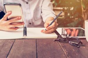 asian woman hand holding phone and pencil writing notebook in coffee shop with vintage toned photo