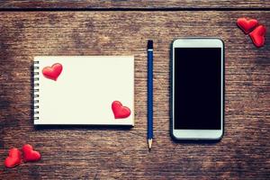 Empty notebook, pencil and red heart on wood table with phone blank copy space screen for your advertising. Valentine background. photo