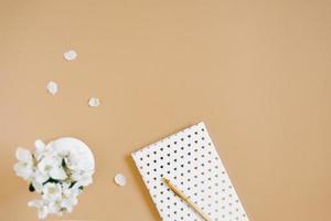 A white notebook with a gold pen on a beige table and flowers in a defocus vase. Blogger's workplace. Top view, copy space photo