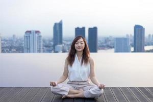 mujer relajantemente practicando meditación a el nadando piscina techo con el ver de urbano horizonte edificio a alcanzar felicidad desde interior paz sabiduría concepto foto