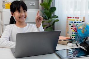 colegiala asiática haciendo su tarea con una laptop en casa. los niños usan aparatos para estudiar. educación y aprendizaje a distancia para niños. educación en el hogar durante la cuarentena. Quédate en casa foto