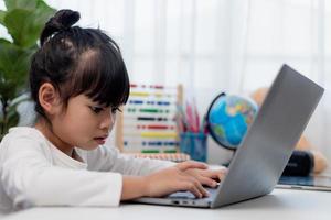 Asian schoolgirl doing her homework with laptop at home. Children use gadgets to study. Education and distance learning for kids. Homeschooling during quarantine. Stay at home photo