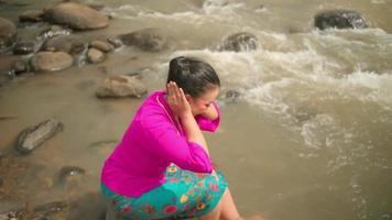 Indonesian woman washes her hands in the river while wearing a pink dress and sitting on the rock inside the village video