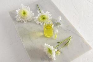 an open bottle of cosmetic natural remedy and a filled eyedropper on a white marble podium and a white background with spring flowers. self-care. photo