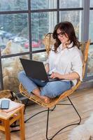Beautiful businesswoman working on a laptop in a chair near the window. Female freelancer connecting to internet via computer. Blogger or journalist writing new article. photo