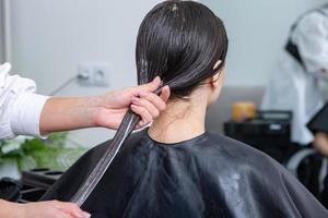 el peluquero aplica una máscara para el cabello al cabello negro liso. cuidado del cabello en el salón de belleza. foto