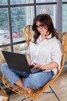 Beautiful businesswoman working on a laptop in a chair near the window. Female freelancer connecting to internet via computer. Blogger or journalist writing new article. photo