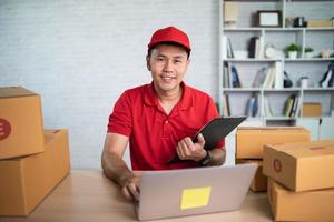 Asian delivery man worker smiling in red uniform work at home , checking list parcel boxes for sending or conveying parcels by mail. Delivery transport concept. photo