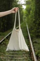 white tote bag canvas fabric with handle mock up design. Close up of woman's hand holding eco or reusable shopping bag on green outdoor background. No plastic bag and ecology concept. photo