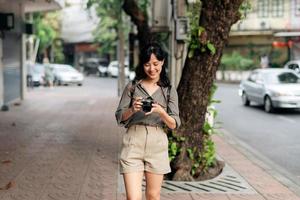joven asiático mujer mochila viajero utilizando digital compacto cámara, disfrutando calle cultural local sitio y sonrisa. foto