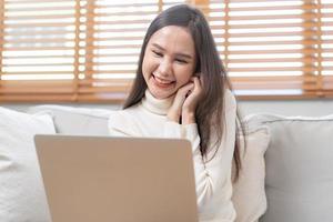 contento alegre, satisfecho asiático joven persona de libre dedicación mujer, niña sentado en sofá o sofá, utilizando ordenador portátil computadora en vivo habitación a hogar, mirando y sonriente a pantalla, empresario disfrutando con trabajar, wfh foto