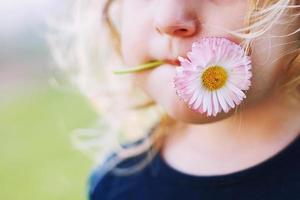 niña con un margarita flor en su boca en un verde antecedentes cerca. labios con flor en césped antecedentes. retrato de un pequeño niña foto