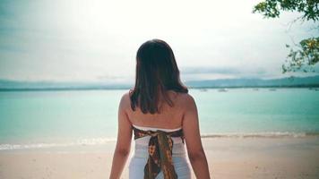 un asiatique femme avec blond cheveux permanent sur le plage tandis que profiter le vue de le bleu mer l'eau et blanc le sable video