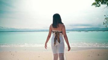 un asiatique femme avec blond cheveux permanent sur le plage tandis que profiter le vue de le bleu mer l'eau et blanc le sable video
