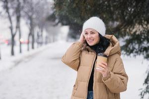 A cute girl holds a disposable cup of coffee and adjusts her hat in winter photo