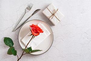 Plate, cutlery, gift and fresh red rose on a textured gray background. Eco holiday serving. Top view. photo