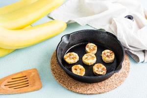 Fried banana slices in a frying pan and fruits nearby on the table. Homemade sweet snacks photo