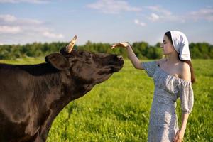 un joven niña en un vaca pasto soportes en un vestir y un pañuelo y golpes un vaca foto