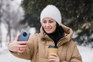 Cute girl laughing and making a video call outside in winter. Hold a disposable cup of coffee photo