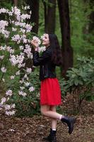 A young girl in a black jacket and a red dress stands near a white rhododendron photo