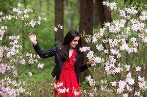 un joven niña en un negro chaqueta y un rojo vestir soportes cerca un blanco rododendro foto