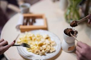 niños comer frutas y postres, bebida té a hogar en el noche cocina. cerca arriba mano con chocolate en un palo para derritiendo. foto