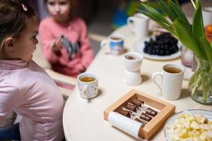 Children eat fruits and desserts, drink tea at home in the evening kitchen. Chocolate on a stick for melting. photo