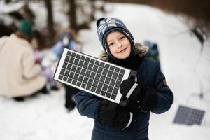 chico con solar panel batería a mano en contra su familia en invierno bosque gasto hora juntos en un picnic. foto