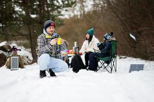 Man with phone at hand against his family in winter forest spending time together on a picnic. photo