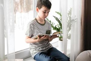 un de seis años chico es leyendo un libro mientras sentado por el ventana. hogar enseñanza foto