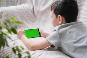 The boy looks at the screen of the phone with a chrome key indoors. An unrecognizable child uses a smartphone with a green screen to watch a video. photo