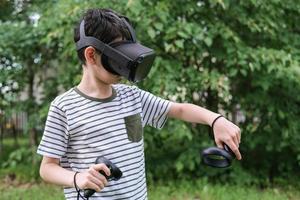 A seven-year-old boy in virtual reality glasses plays outside in the park. Children using technology photo