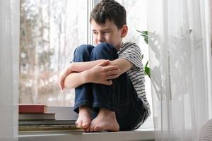 A sad boy sits on the windowsill hugging his knees. Bad mood, depression. The boy is sad alone at home photo